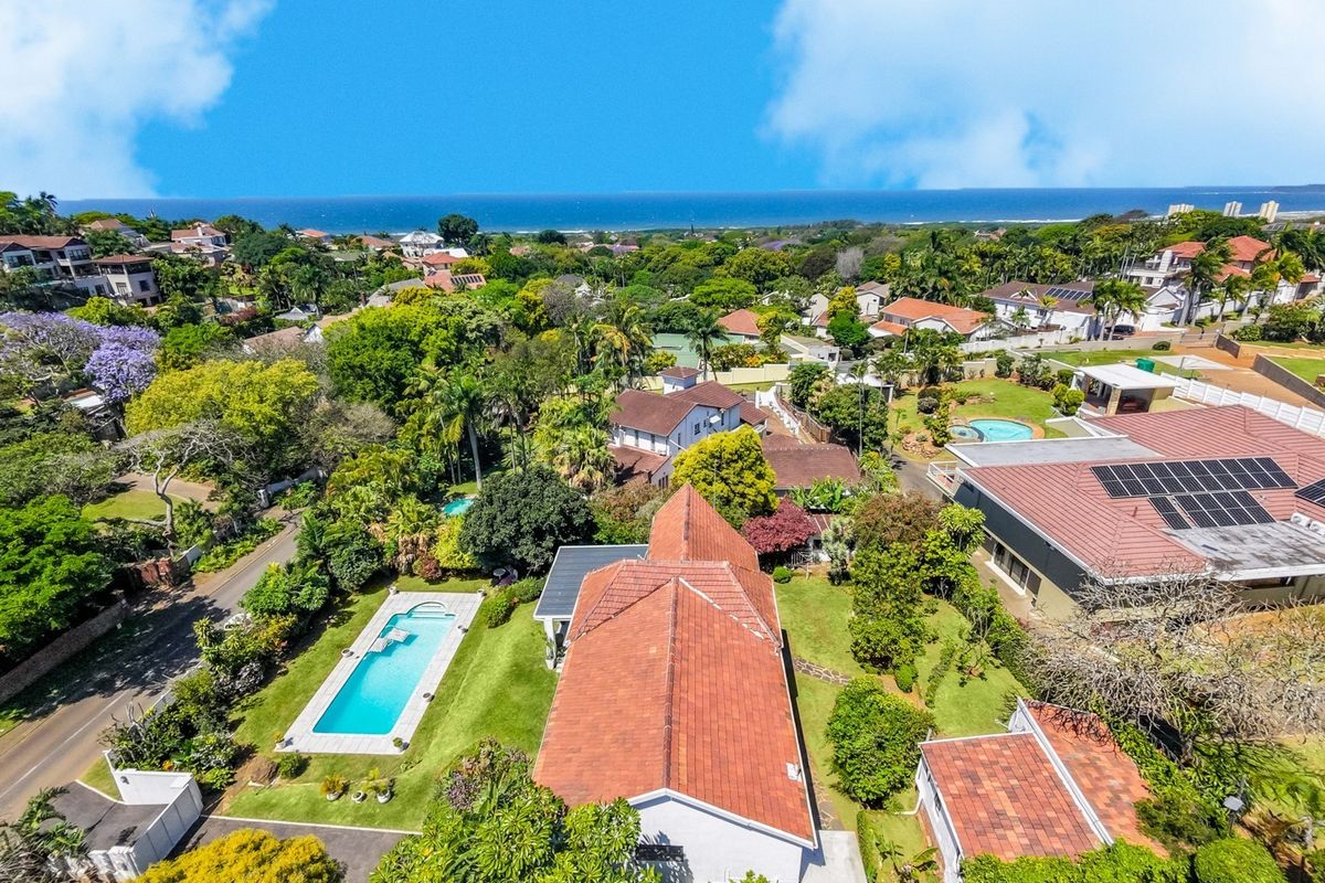 Aerial view of the property showcasing the swimming pool, tiled roof with solar panels, and surrounding landscaped garden in a coastal neighborhood.