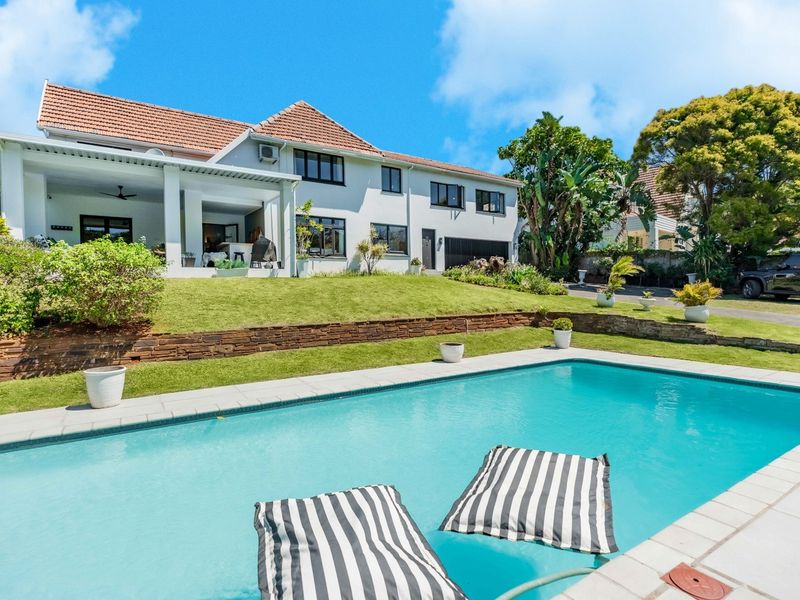 Rear view of the property featuring a swimming pool, a covered patio area, and a well-maintained garden with stone retaining walls.