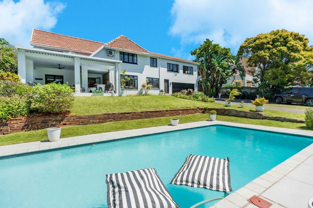 Rear view of the property featuring a swimming pool, a covered patio area, and a well-maintained garden with stone retaining walls.