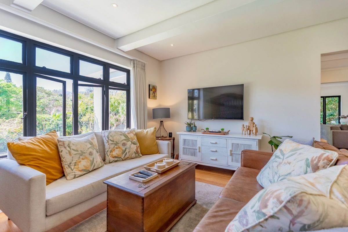 A bright and well-maintained living room featuring hardwood flooring, recessed lighting, and large black-framed windows overlooking a lush garden.