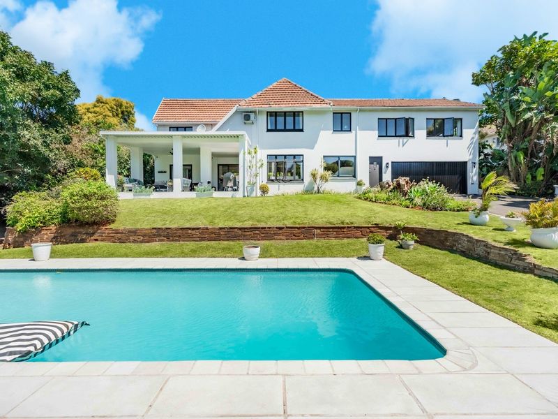 Rear view of a well-maintained two-story house featuring a swimming pool, covered patio, and landscaped garden with stone retaining walls.