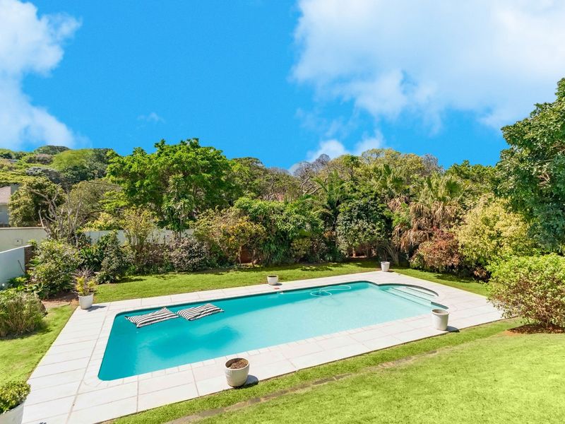 A well-maintained rectangular swimming pool set within a lush, landscaped garden with mature trees and a paved pool surround.