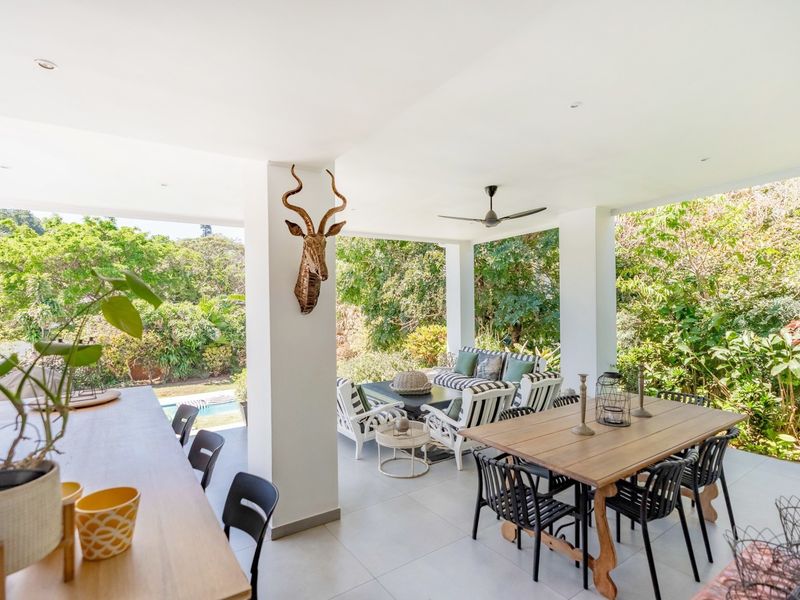 A spacious covered patio featuring tiled flooring, recessed lighting, and a ceiling fan, overlooking a lush garden and pool area.