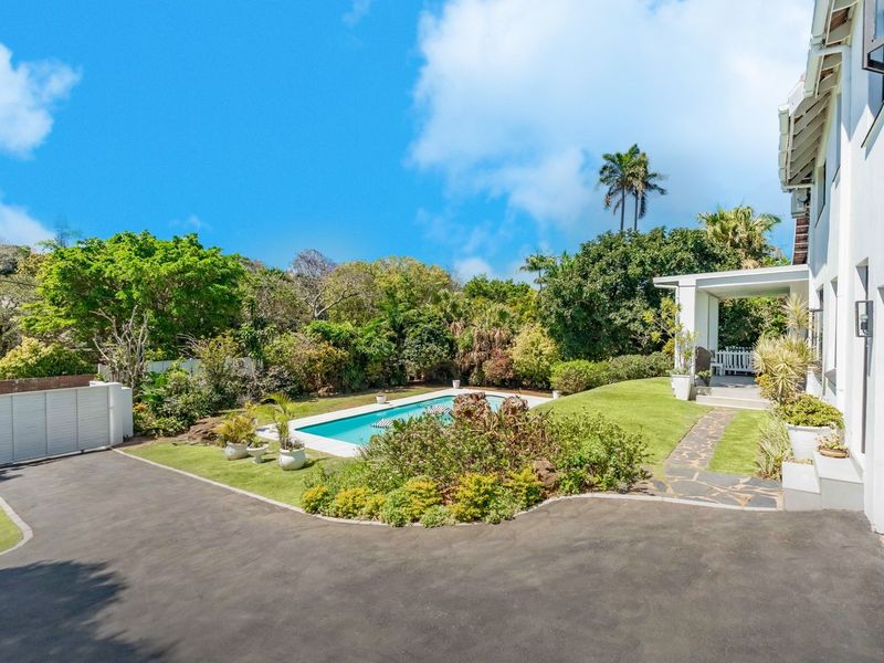 A well-maintained rear garden featuring a swimming pool, paved driveway, stone pathway, and a covered patio area adjacent to the main house.