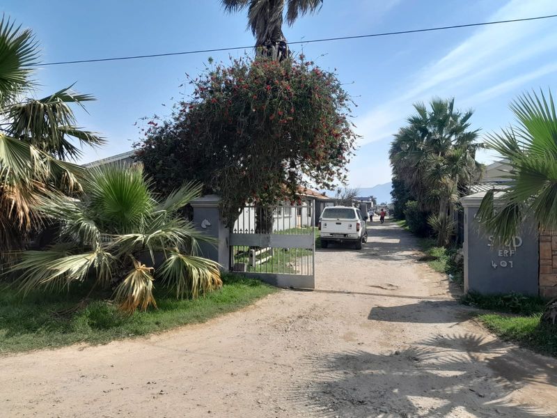 Front entrance view of the property featuring a metal gate, masonry pillars, and an unpaved driveway surrounded by established palm trees.