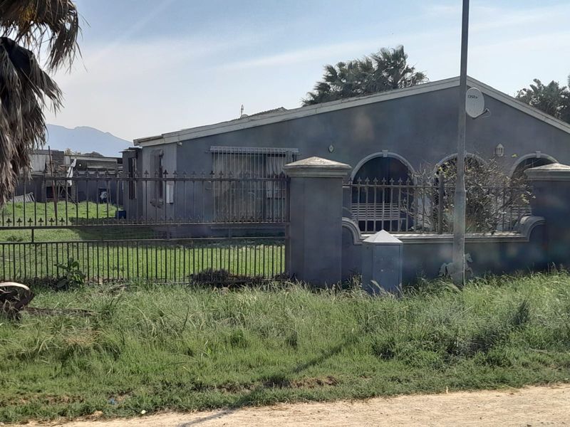 Front exterior view of a single-story house featuring arched window openings, metal perimeter fencing, and masonry pillars, set on a grassed plot.
