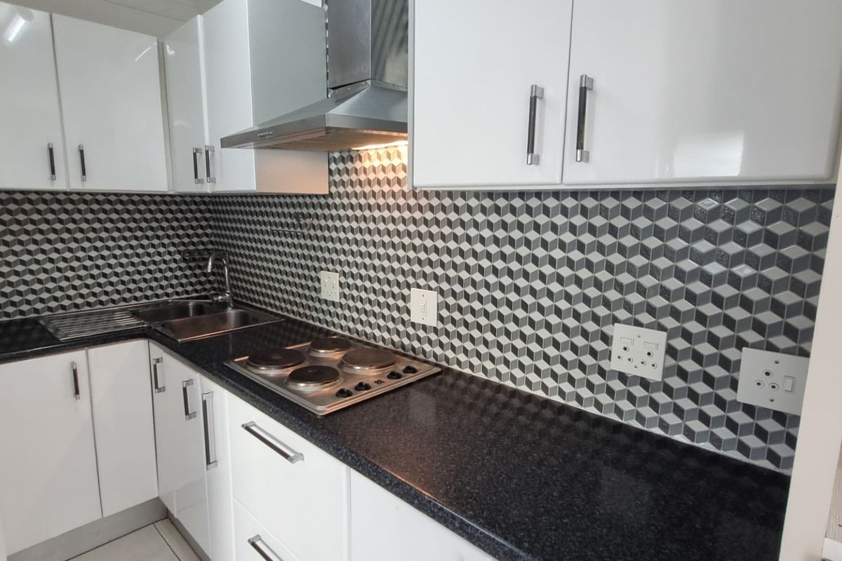 A modern, compact kitchen featuring white cabinetry, black granite countertops, a stainless steel hob with extractor fan, and a striking geometric tile backsplash.