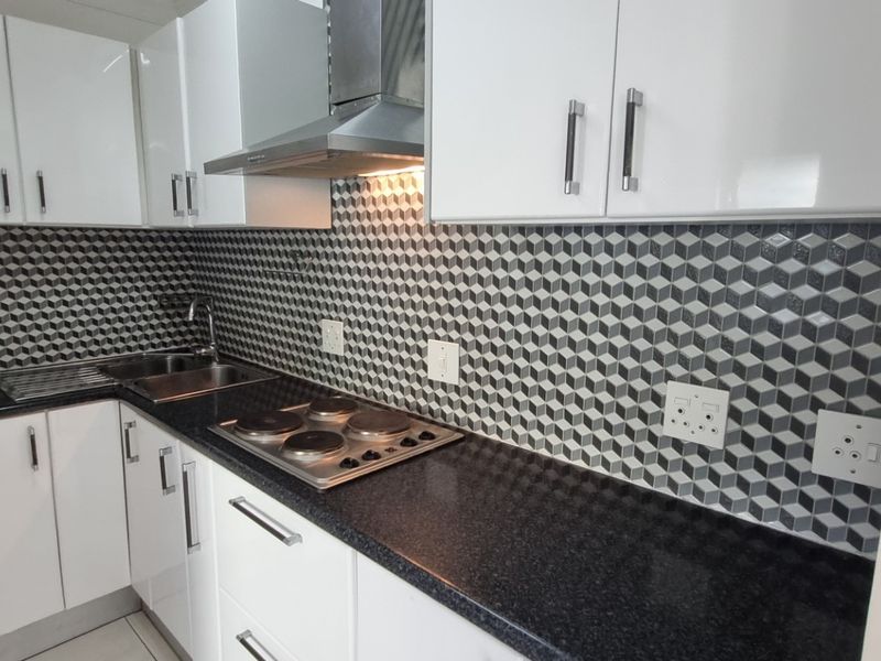 A modern, compact kitchen featuring white cabinetry, black granite countertops, a stainless steel hob with extractor fan, and a striking geometric tile backsplash.