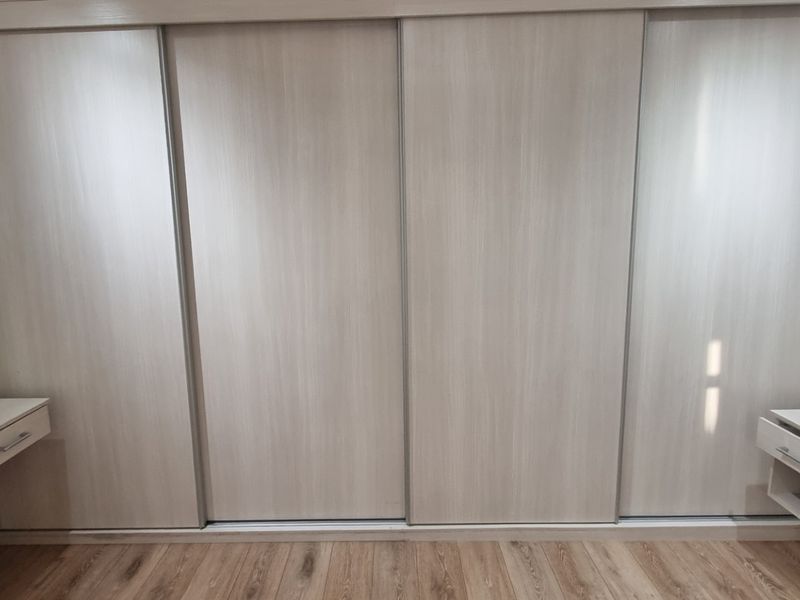 A clean bedroom featuring a large built-in sliding door wardrobe and matching floating vanity units with modern laminate flooring.