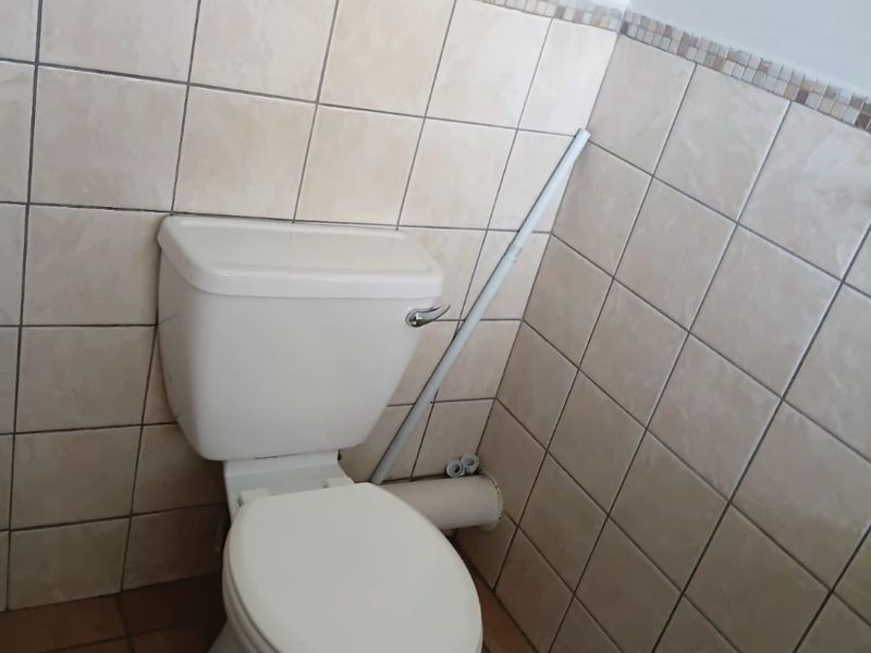 A compact bathroom featuring ceramic wall and floor tiling, a standard toilet, and a glass shelf, currently showing signs of dated finishes and exposed plumbing.