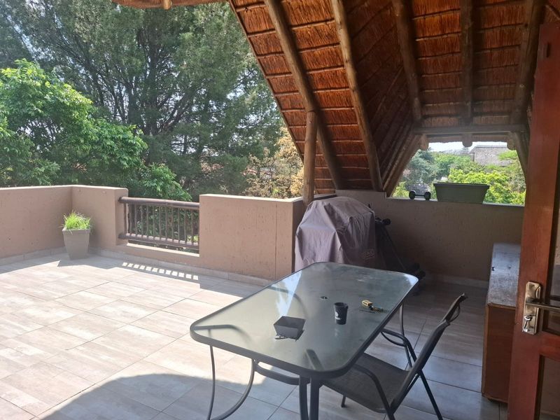 A spacious covered balcony featuring a traditional thatched roof, tiled flooring, and a masonry parapet wall with railing, overlooking mature trees.