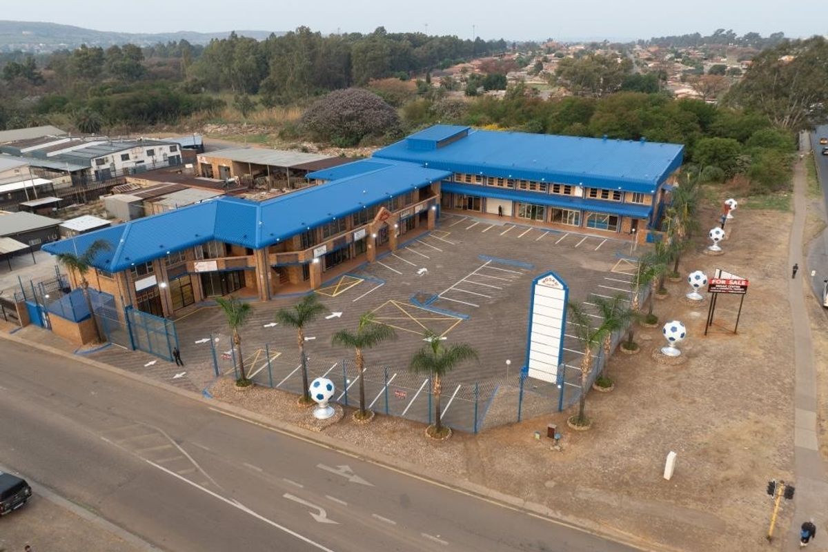 Aerial view of a commercial property featuring a blue metal roof, paved parking lot, perimeter fencing, and prominent signage pylon.