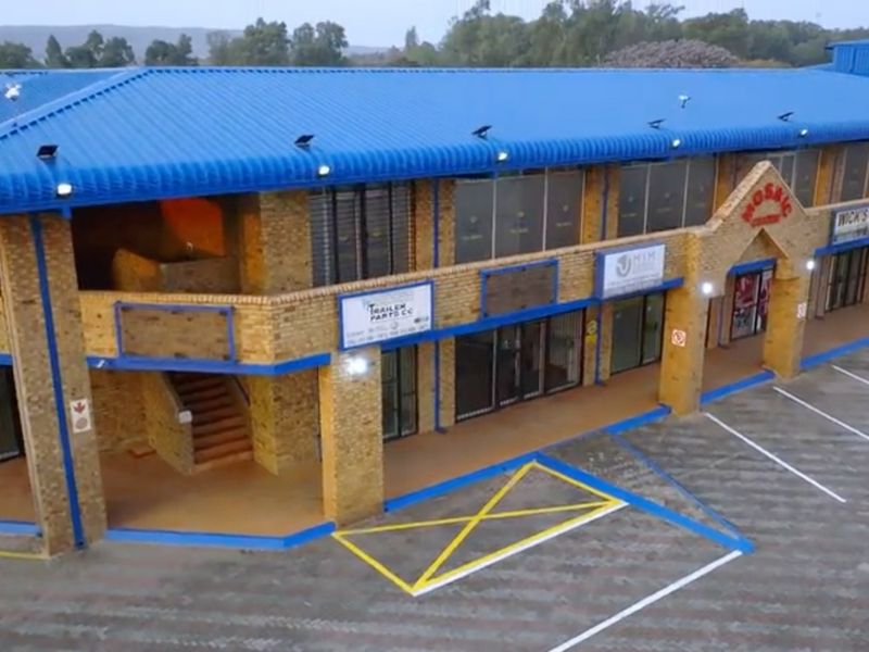 Aerial view of a commercial retail complex featuring a blue corrugated roof, brick facade, and dedicated paved parking area with multiple storefront entrances.