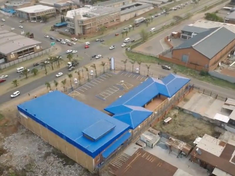 Aerial view of a commercial property featuring a blue metal roof, dedicated parking area, and perimeter fencing, situated near a busy intersection.