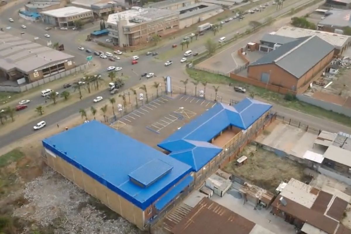 Aerial view of a commercial property featuring a blue metal roof, dedicated parking area, and perimeter fencing, situated near a busy intersection.