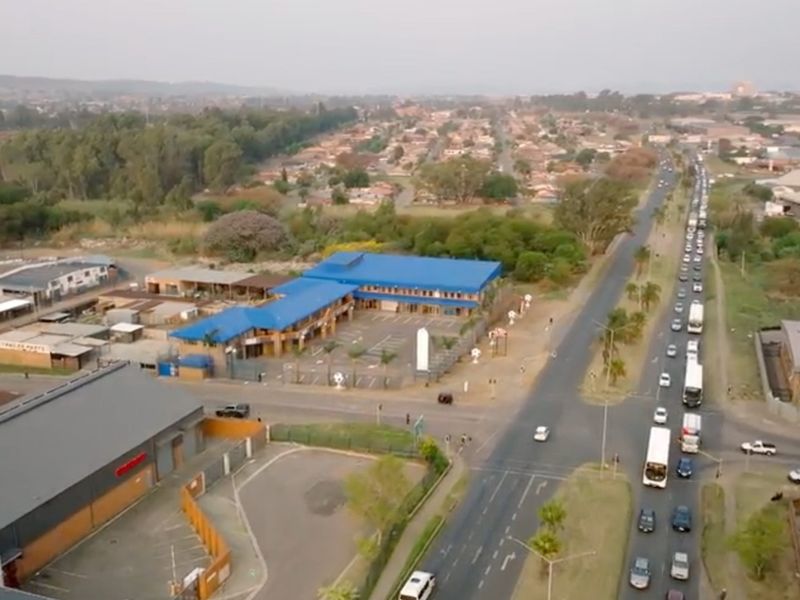 Aerial view of a commercial property complex featuring a blue metal roof, paved parking area, and perimeter fencing situated near a busy road intersection.