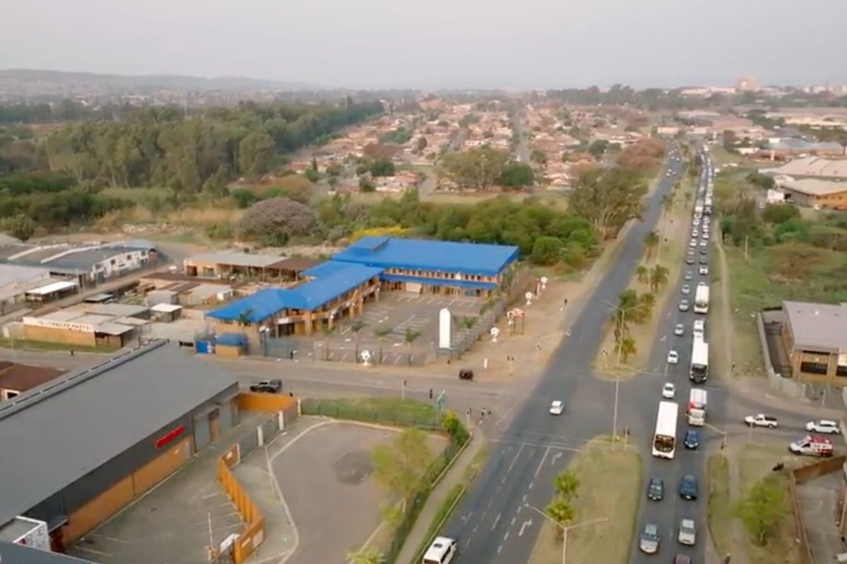 Aerial view of a commercial property complex featuring a blue metal roof, paved parking area, and perimeter fencing situated near a busy road intersection.