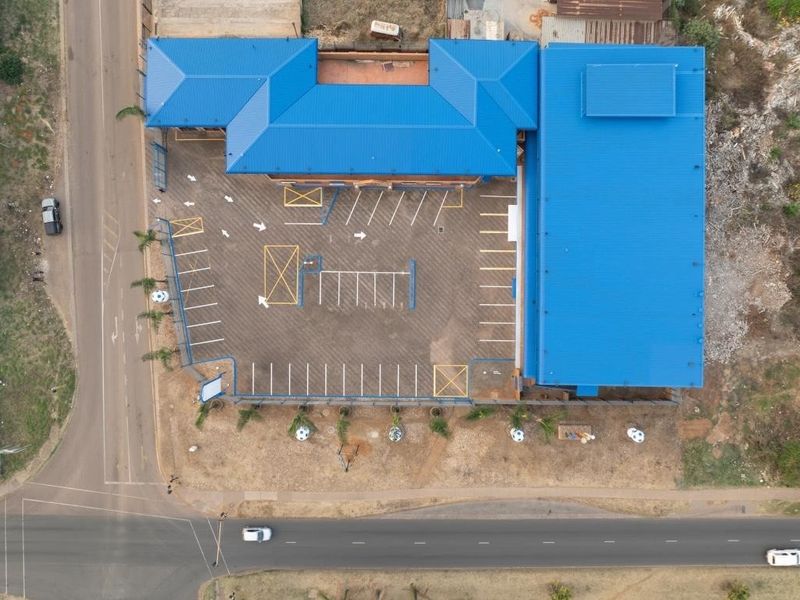 Aerial view of a commercial property featuring a large metal-roofed building, an extensive asphalt parking area with marked bays, and perimeter fencing.