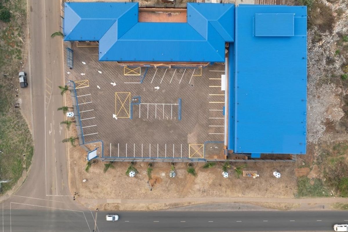 Aerial view of a commercial property featuring a large metal-roofed building, an extensive asphalt parking area with marked bays, and perimeter fencing.