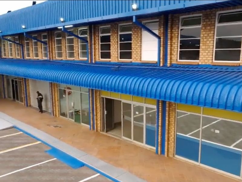 Exterior view of a commercial building featuring a blue corrugated roof, brick facade, and multiple glass storefront entrances with adjacent paved parking.