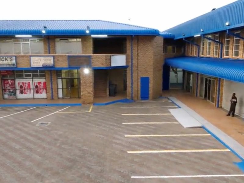 An aerial view of a commercial retail complex featuring brick facades, blue corrugated roofing, and a paved parking area with multiple storefront entrances.