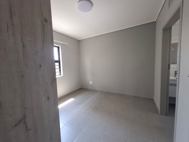 A clean, empty bedroom featuring neutral tiled flooring, a window with security bars, and a built-in cupboard, with an adjacent bathroom visible.