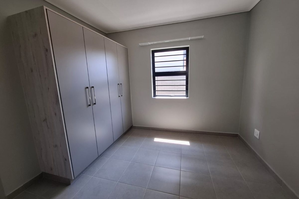 A clean, empty bedroom featuring neutral tiled flooring, built-in cupboards, and a window fitted with security bars.