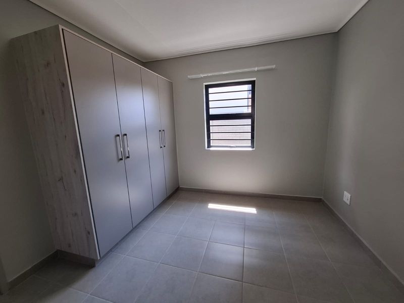 A clean, empty bedroom featuring neutral tiled flooring, built-in cupboards, and a window fitted with security bars.