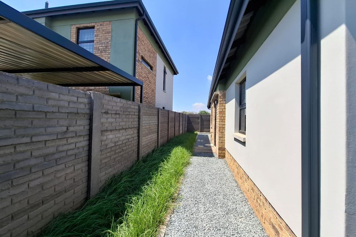 A side exterior view showing a gravel pathway between a brick boundary wall and the house structure, featuring a carport and established landscaping.