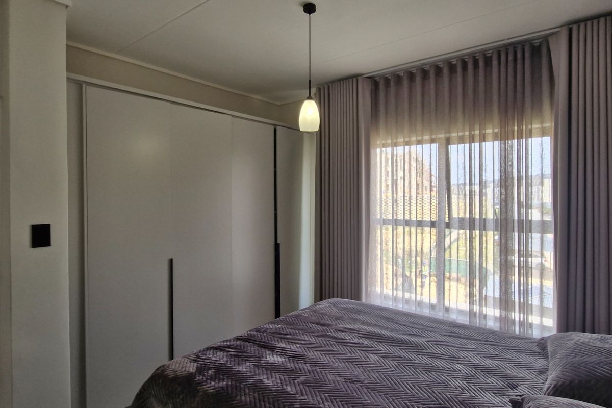 A well-maintained bedroom featuring a large built-in wardrobe, modern pendant lighting, and floor-to-ceiling curtains over a sliding window.