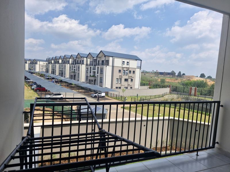 A covered balcony with tiled flooring and a metal balustrade, offering an elevated view of the apartment complex and surrounding grounds.