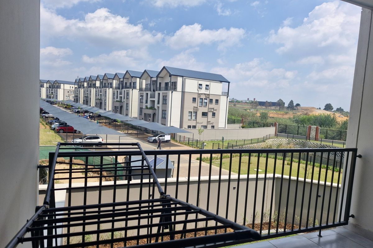 A covered balcony with tiled flooring and a metal balustrade, offering an elevated view of the apartment complex and surrounding grounds.