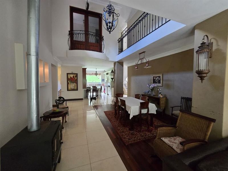 Spacious open-plan living and dining area featuring a double-volume ceiling, mezzanine level, tiled and hardwood flooring, and a prominent freestanding fireplace.