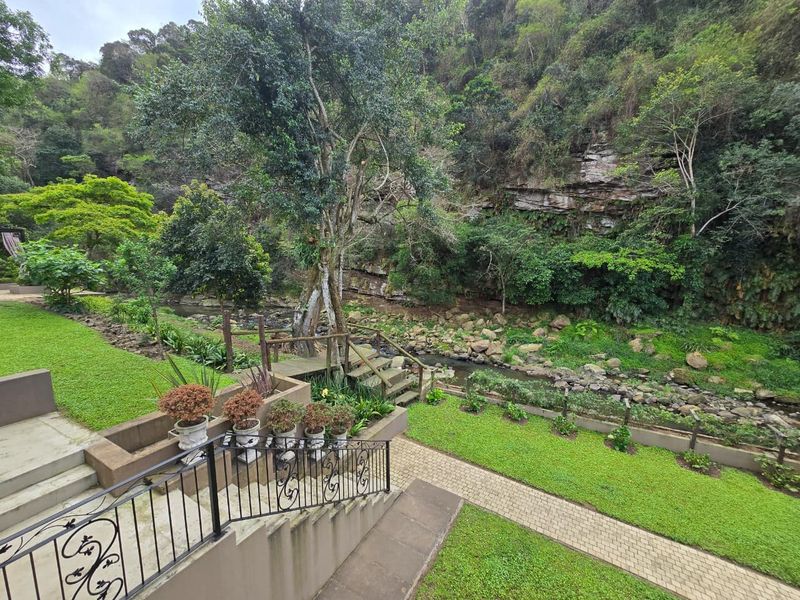 A beautifully landscaped garden featuring a paved walkway, concrete stairs with wrought iron railings, and a natural stream set against a rocky cliff backdrop.