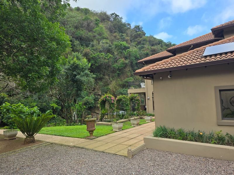Rear view of the property featuring a tiled roof, solar panel, paved walkway, and a well-maintained garden area set against a natural hillside backdrop.