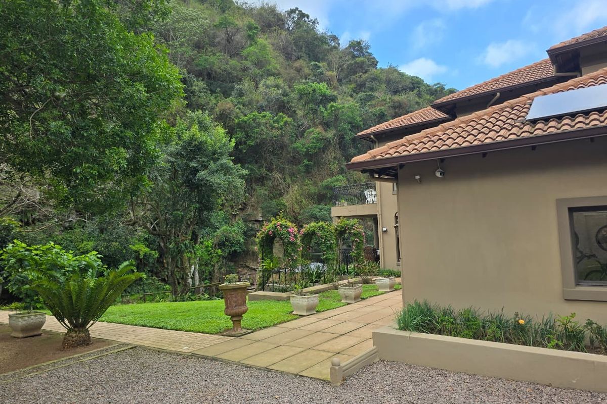 Rear view of the property featuring a tiled roof, solar panel, paved walkway, and a well-maintained garden area set against a natural hillside backdrop.