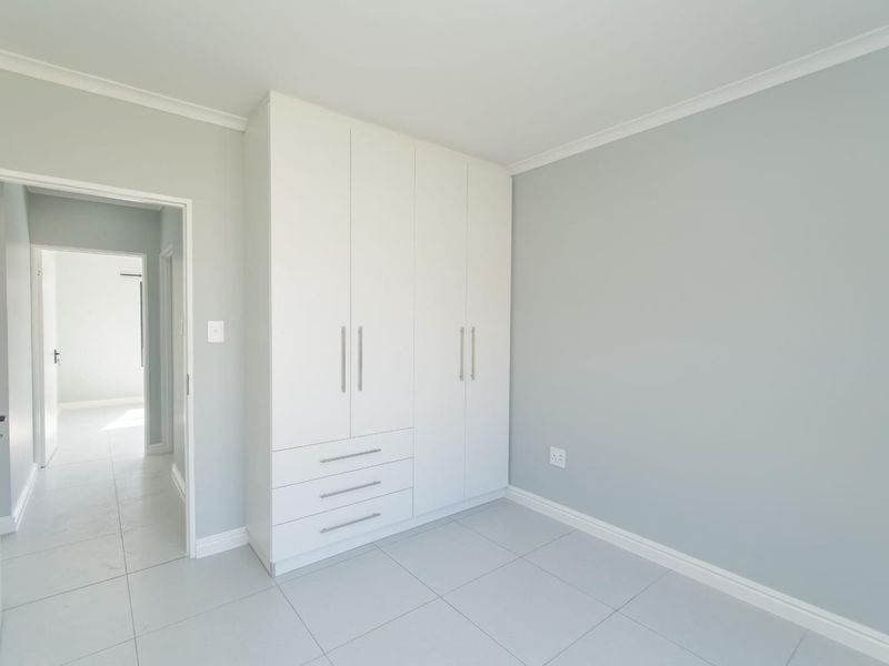A bright, empty bedroom featuring neutral grey walls, white tiled flooring, and a modern built-in wardrobe with integrated drawers.