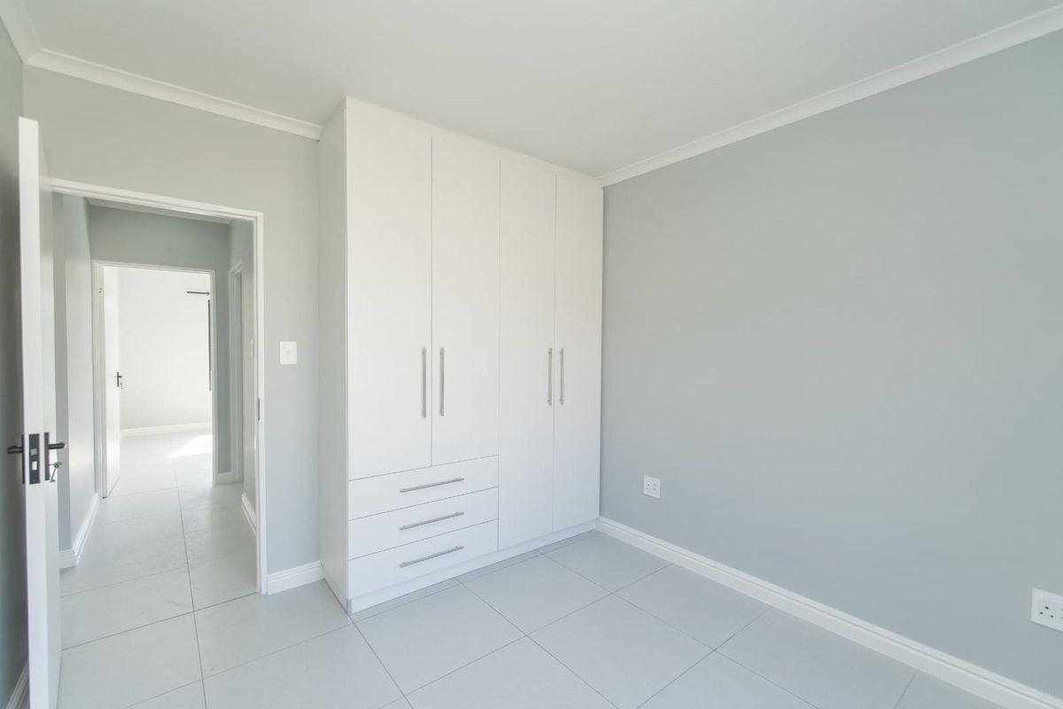 A bright, empty bedroom featuring neutral grey walls, white tiled flooring, and a modern built-in wardrobe with integrated drawers.