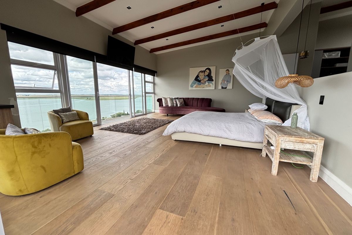 Spacious bedroom featuring exposed timber ceiling beams, engineered wood flooring, and expansive sliding glass doors offering panoramic water views.