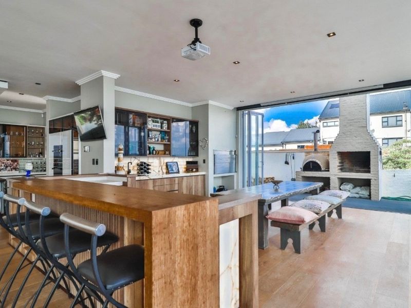 Spacious open-plan entertainment area featuring a custom built-in bar, recessed lighting, and seamless transition to an outdoor patio with a brick fireplace and pizza oven.