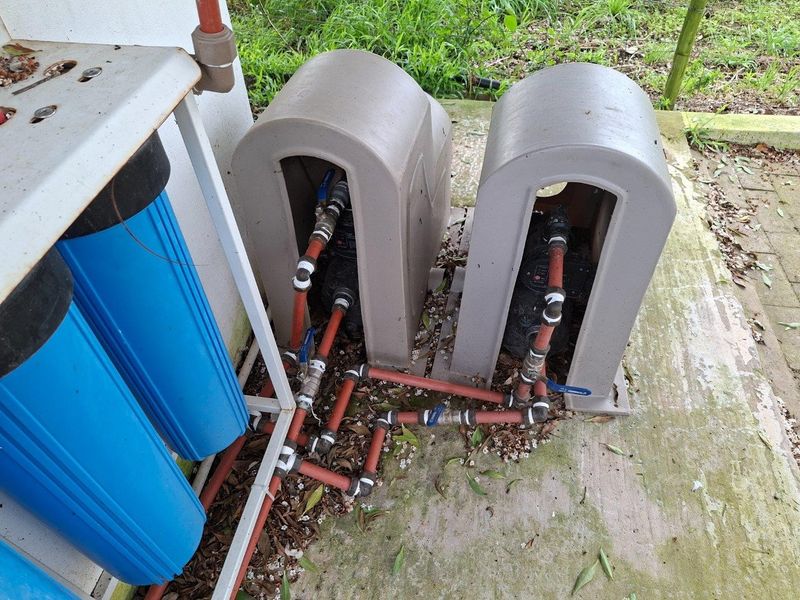 Outdoor utility area featuring a dual water pump system with protective covers and a multi-stage water filtration unit connected via external plumbing.