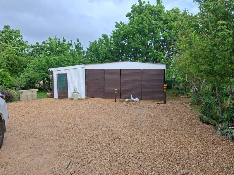 A detached garage structure with corrugated metal siding and double garage doors, situated on a gravel driveway within a garden setting.