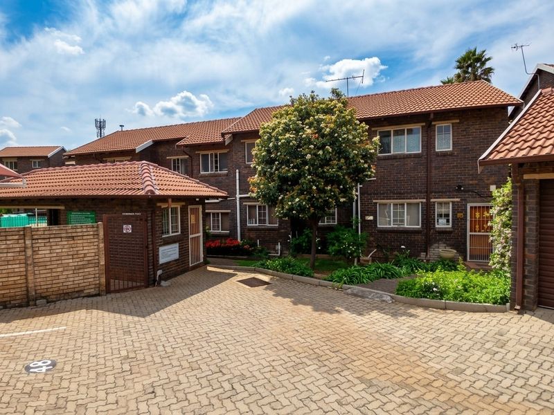 Front exterior view of a brick townhouse complex featuring a paved driveway, tiled roofing, and established garden beds.