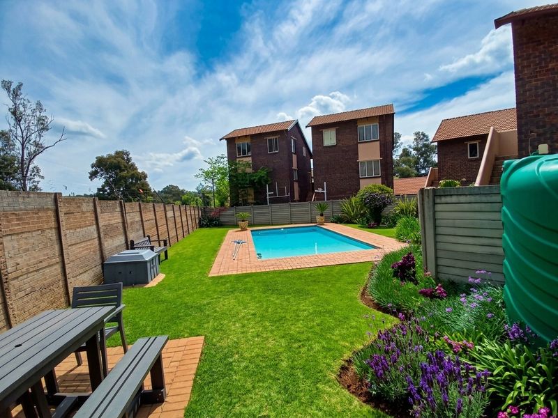 A well-maintained outdoor pool area featuring a swimming pool, brick paving, landscaped garden, and a water tank, set within a secure townhouse complex.