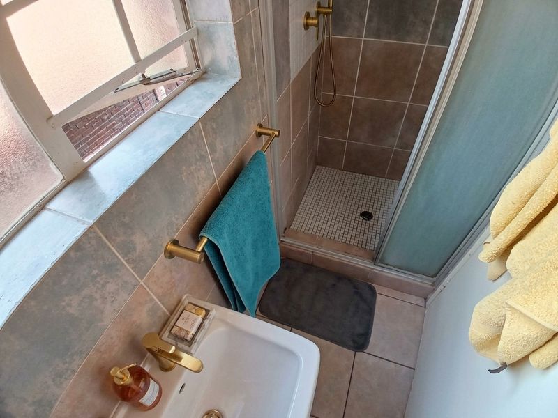 A compact bathroom featuring a tiled shower enclosure, ceramic basin with brass fixtures, and neutral wall tiling.