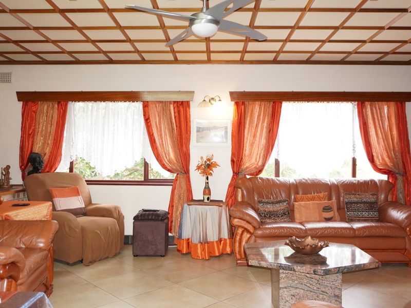A spacious living room featuring a distinctive coffered ceiling, ceiling fan, tiled flooring, and an exposed brick feature wall.
