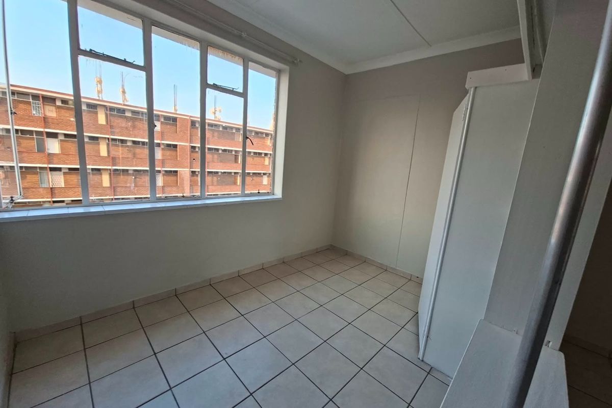 A bright, empty bedroom featuring tiled flooring, a large window providing natural light, and a built-in cupboard for storage.