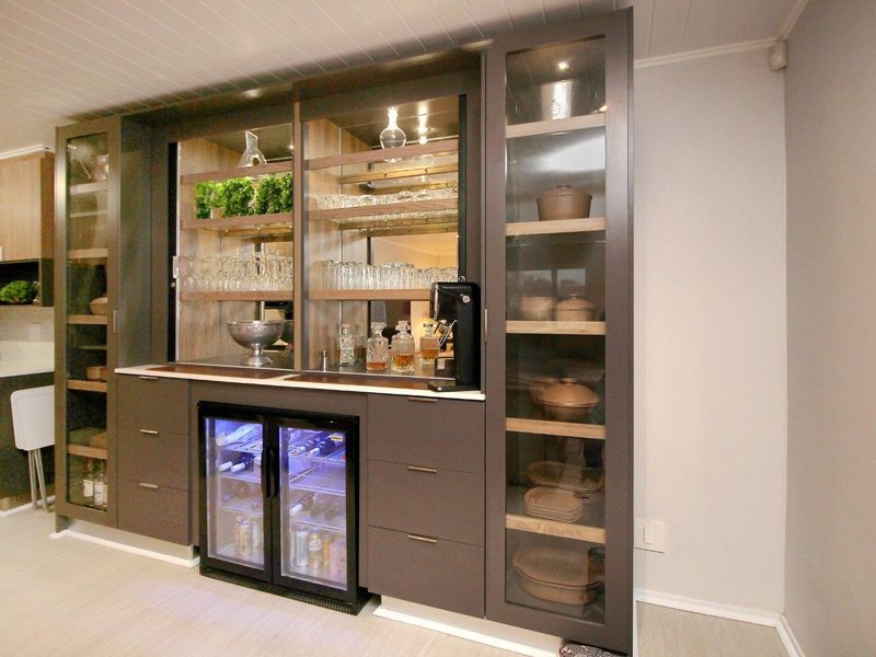 A modern, well-appointed built-in home bar featuring dark cabinetry, glass display shelving, a mirrored backsplash, and an integrated wine cooler.
