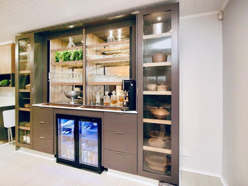 A sophisticated built-in home bar featuring custom cabinetry, glass-fronted display shelving, a mirrored backsplash, and an integrated wine cooler.