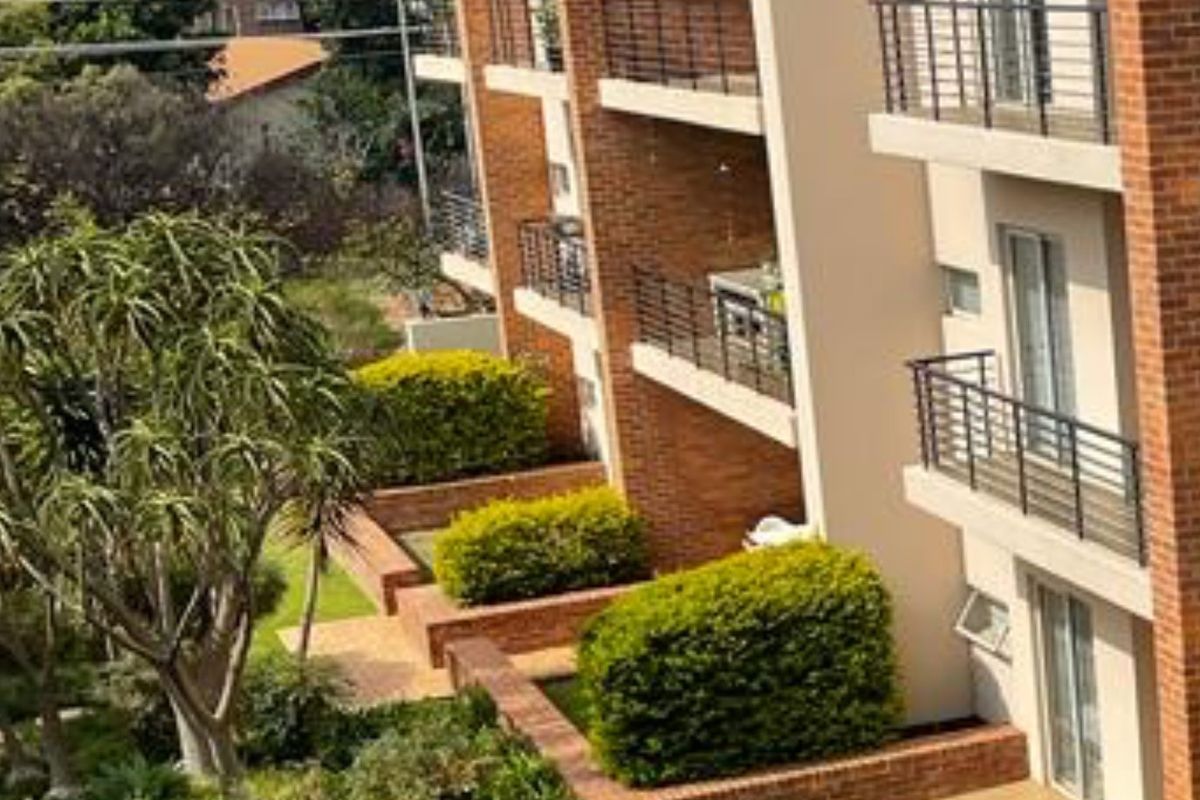 Exterior view of an apartment building featuring brick facade, multiple balconies with metal railings, and well-maintained landscaped garden areas.